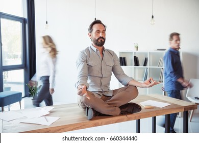 Relaxed businessman meditating in lotus position while coworkers moving in office
