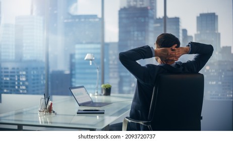 Relaxed Businessman In A Suit Sitting At A Desk In Modern Office, Using Laptop Computer, Looking At Window City View. Successful Finance Manager Stretching At His Desk While Planning Work Projects.