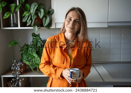 Relaxed calm blond woman holding mug standing in kitchen surrounded by houseplants. Portrait of smiling confident female in orange dress holds tea cup looking at camera. Pretty Scandinavian girl. 