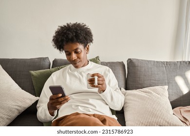 Relaxed calm gen z African American teen, ethnic guy sitting on couch at home chilling holding cellphone, using mobile apps checking social media on cell phone drinking coffee. Authentic shot. - Powered by Shutterstock