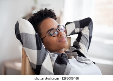 Relaxed calm happy black woman resting taking healthy break holding hands behind head breathing fresh air in office, tired african girl enjoying peaceful mood stress relief dreaming lounging concept