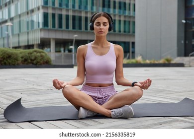 Relaxed calm woman sits in lotus pose on fitness mat meditates and listens pleasant music via headphones practices yoga in urban setting wears sports clothes keeps eyes closed breathes deeply - Powered by Shutterstock