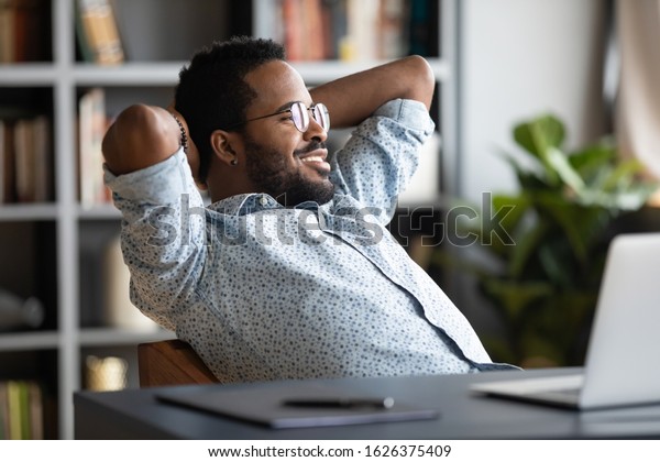 Relaxed calm young african businessman resting\
looking away sit at desk with laptop hands behind head, satisfied\
office employee take break feel stress relief peace of mind concept\
chill at work