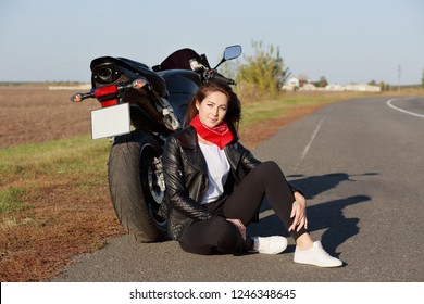 Relaxed Carefree Female Motorcyclist Wears Black Clothes And White Trainers, Sits At Asphalt Near Motorbike, Enjoys Adventure And Racing In Countryside, Prepares For Motorcross. Leisure Concept