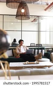 Relaxed Casually Dressed Young Man With Mobile Phone And Wireless Headphones Working On Laptop In Seating Area Of Modern Open Plan Office