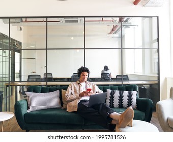 Relaxed Casually Dressed Young Man With Mobile Phone And Wireless Headphones Working On Laptop In Seating Area Of Modern Open Plan Office