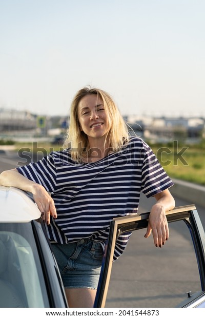 Relaxed caucasian female standing at car open\
door at parking. Happy woman vehicle owner wear casual t-shirt and\
jeans shirts ready for vacation by auto road trip. Confident girl\
driver at automobile