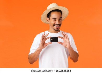 Relaxed And Cheeky Cute African-american Man In White T-shirt, Summer Hat, Wink Camera Assured And Pleased, Recommend Bank Service, Holding Credit Card, Standing Orange Background