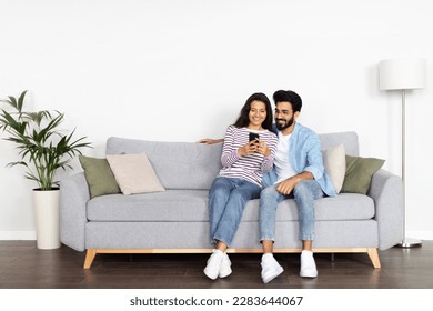 Relaxed cheerful beautiful young multicultural couple middle eastern guy and indian woman sitting on sofa at home, using smartphone and smiling, shopping online, surfing on Internet, copy space - Powered by Shutterstock