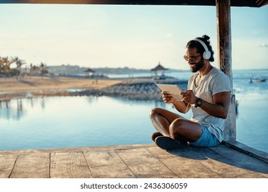 Relaxed and cheerful. Work and vacation. Outdoor portrait of happy young african man using tablet computer on deck near the sea. - Powered by Shutterstock