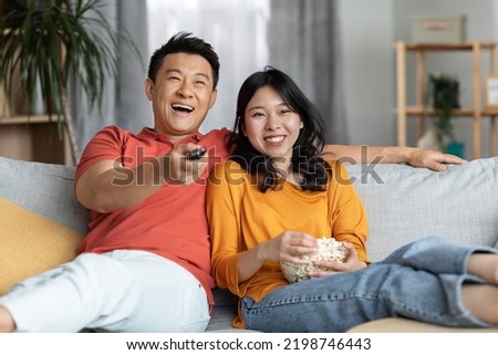 Relaxed chinese family watching TV together at home, happy husband and wife sitting on couch in living room, eating popcorn and laughing, man holding remote controller, copy space