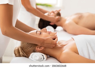 Relaxed Couple Enjoying Massage Lying Together On Beds, Relaxing While Masseurs Massaging Their Neck And Face At Spa Center Indoors. Relaxation, Beauty And Wellness Concept. Selective Focus