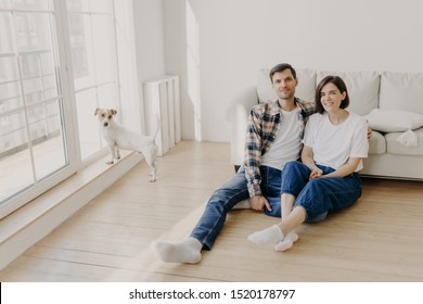 Relaxed Couple Sit On Floor Near Couch, Embrace And Smile, Dressed In Casual Clothes And White Socks, Enjoy Domestic Atmosphere, Their Dog Stands Near Balcony Window In Empty Spacious Living Room