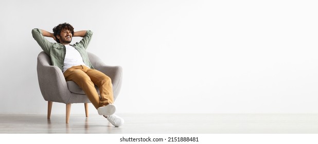 Relaxed curly bearded dark-skinned guy in casual outfit sitting at armchair with arms behind head, looking at copy space for advertisement or text and smiling over white studio background, panorama - Shutterstock ID 2151888481