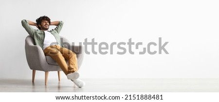 Relaxed curly bearded dark-skinned guy in casual outfit sitting at armchair with arms behind head, looking at copy space for advertisement or text and smiling over white studio background, panorama