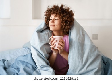 Relaxed curly woman with closed eyes wrapped in blanket while holding mug of hot beverage on bed in weekend