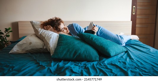 Relaxed curly-haired caucasian woman lying on bed holding book in hands reading wearing nightwear. Lifestyle concept. Education concept. Beautiful female person - Powered by Shutterstock