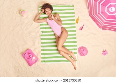 Relaxed dark skinned woman in pink bikini lies on green striped towel has dreamy expression poses on sandy beach with parasol energetic drinks sand toys headphones around. Summer leisure concept - Powered by Shutterstock