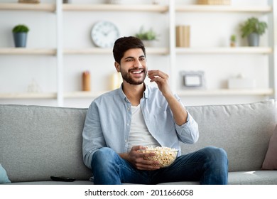 Relaxed Eastern Man Spending Free Time At Home, Eating Popcorn And Watching TV. Excited Arab Guy Enjoying Movie While Relaxing On Couch In Living Room Interior, Copy Space