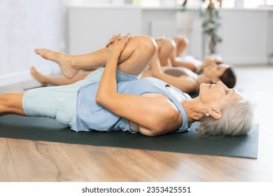 Relaxed elderly woman doing yoga with group of women in studio, performing hamstring and low back stretching Ardha Pawanmuktasana, lying on mat with knee pressed to chest with hands - Powered by Shutterstock