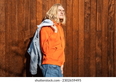 Relaxed European man in denim jacket over shoulder with close eyes stands near wooden wall in sun. Dreamy scandinavian blond guy freelancer enjoy sunny day take break resting outdoor near coworking - Powered by Shutterstock