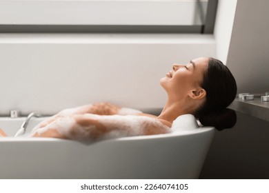 Relaxed Female Taking Bath With Foam Lying In Bathtub In Modern Bathroom Indoors. Woman Bathing Enjoying Bodycare Routine With Eyes Closed At Home. Spa And Wellness. Side View, Selective Focus