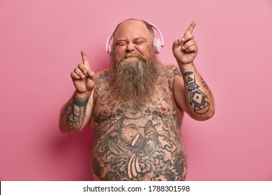 Relaxed funny thick man with naked body, tattooed arms and stomach, dances while listens music, moves arms and closes eyes in enjoyment, wears headphones on ears, has fun and feels aspiration