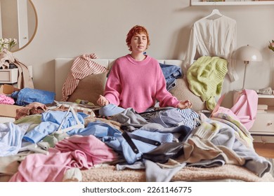 Relaxed ginger young woman meditates on bed breathes deeply sits in lotus pose surrounded by variety of unfitting clothes makes revision of her garment at home collects clothing for donation