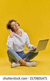 Relaxed Girl Listening To Music On Laptop, Sitting With Crossed Legs On Floor. Front View Of Young Girl With Closed Eyes, Enjoying Melody In Earphones, Isolated On Orange Background. Concept Of Relax.