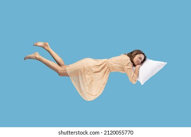 Relaxed girl in yellow dress levitating in mid-air, sleeping on stomach lying comfortable cozy on pillow, keeping eyes closed, watching peaceful dream. indoor studio shot isolated on blue