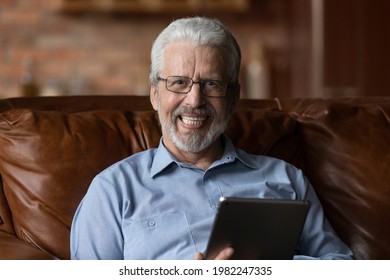 Relaxed Grey-haired Man In Glasses Sit On Sofa At Home With Tablet Device, Head Shot Portrait. Older Generation And Modern Wireless Tech, Easy Usage Of Gadget, Online Services For Aged Citizen Concept