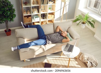 Relaxed Guy Sleeping On Comfortable Beige Couch At Home. Man Taking Break From Work And Napping On Soft Couch With Laptop On Coffee Table Beside In Cosy Modern Living Room. High Angle. Rest Concept