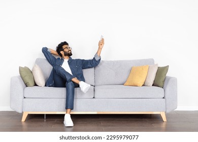 Relaxed handsome bearded middle eastern young man in casual outfit and eyeglasses sitting on couch at home, holding remote control, turning on ac, pointing towards copy space on white wall - Powered by Shutterstock