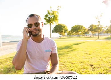 Relaxed Handsome Guy Talking On Cell While Enjoying Leisure Time Outdoors. Latin Man Sitting On Grass In Park, Speaking On Cellphone. Phone Talk Concept