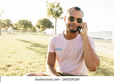 Relaxed Handsome Guy Talking On Cell While Enjoying Leisure Time Outdoors. Latin Man Sitting On Grass In Park, Speaking On Cellphone. Phone Talk Concept