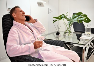 Relaxed Handsome Mature 50 Years Old Man In White Terry Bathrobe Sitting On An Armchair With A Glass Of Fresh Water And Talking On Mobile Phone At Wellness Spa Center