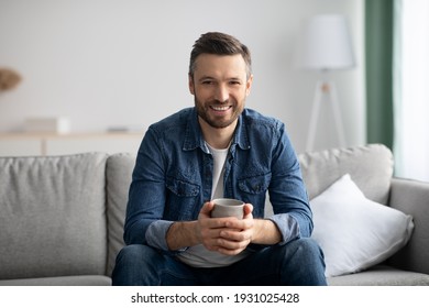 Relaxed handsome middle-aged man in casual outfit sitting on couch with mug of tea, caucasian bearded man drinking coffee in cozy living room at home, smiling at camera, copy space - Powered by Shutterstock