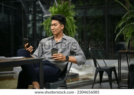 Relaxed and handsome millennial Asian man using his smartphone and sipping coffee at a coffee shop outdoor space.