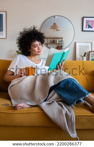 Relaxed, happy African american woman at home living room sitting on the couch, reading a book and drinking tea