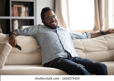 Relaxed Happy Afro American Young Man Sit On Comfortable Sofa Hold Remote Control Watching Tv Concept, Millennial African Guy Lounge In Living Room Spend Weekend Enjoying Television Alone At Home