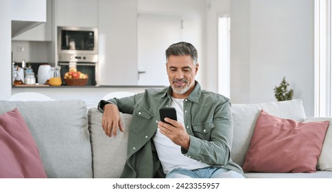 Relaxed happy older mature middle aged man holding mobile phone using cellphone sitting at home on sofa in modern living room, chatting online, texting messages, reading news, shopping in apps. - Powered by Shutterstock