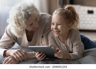 Relaxed happy older senior grandmother having fun with laughing sincere cute grandchild girl, using digital touchpad gadget lying together on soft pillows on floor, modern technology addiction concept - Powered by Shutterstock