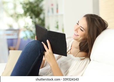 Relaxed Happy Woman Reading A Book In An Ebook Reader Sitting On A Couch At Home