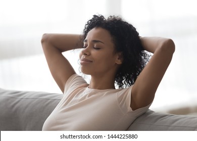 Relaxed happy young african woman resting lounging meditating sit on sofa with eyes closed hands behind head, healthy lazy black girl breathing fresh air sit on couch at home enjoy no stress free day