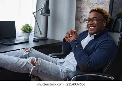 Relaxed, Happy Young African-American Man In Ripped Jeans Put His Feet On The Table. Black Guy In Loose Clothes, An Earring In His Ear And An African Haircut Smiles Looking At The Camera.