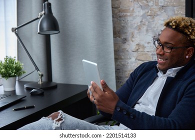 Relaxed, Happy Young African-American In Ripped Jeans Put His Feet On The Table. A Black Guy In Loose Clothes, With An Earring In His Ear And An African Haircut Smiles Using A Digital Tablet