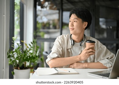 Relaxed and happy young Asian male college student or freelancer remote working at the coffee shop, sipping coffee, pensively thinking something while looking outside of the coffee shop. - Powered by Shutterstock