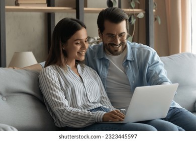 Relaxed happy young family couple sitting on sofa with computer, enjoying watching funny movie, web surfing information, planning weekend or vacation, spending time online, modern tech addiction.
