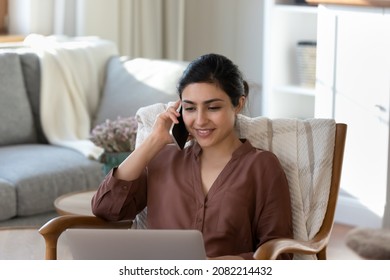 Relaxed happy young indian woman holding phone call conversation, sitting in cozy armchair with computer on laps, making order in internet store, communicating with friends, multitasking at home. - Powered by Shutterstock