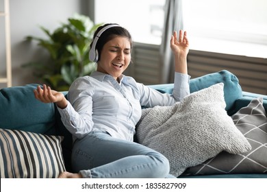 Relaxed indian ethnicity young woman in headphones lean on cozy sofa, enjoying favorite music at home, happy girl singing song, moving in rhythm seated on sofa. Lifestyle, free time and hobby concept